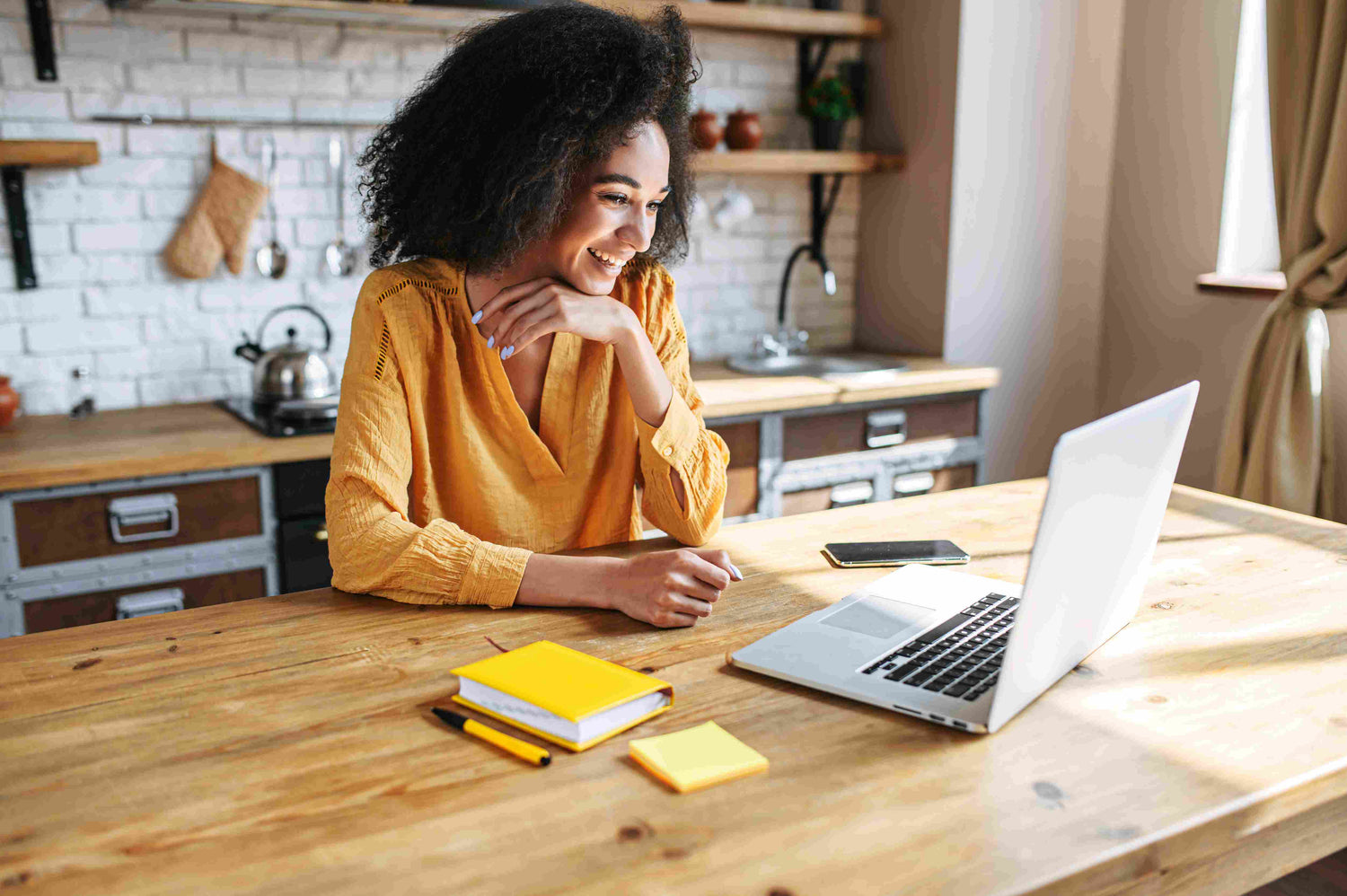 A woman in her kitchen standing looking at her laptop smiling with a notebook next to her