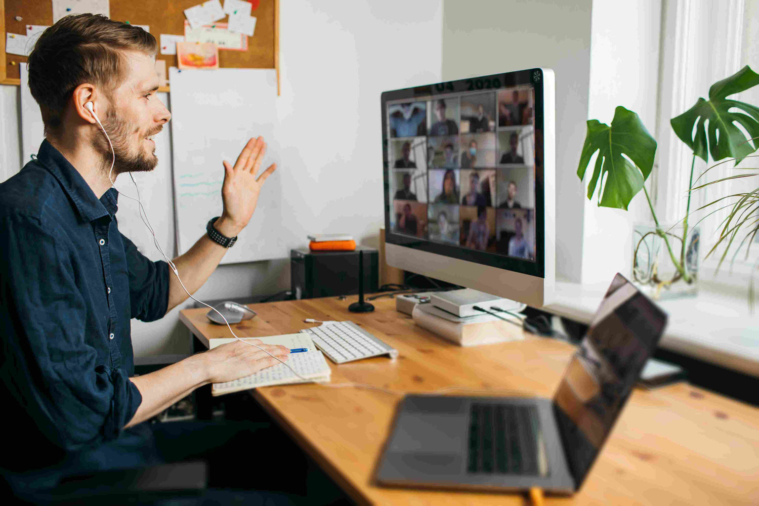 A man sitting at a desk in his home office waiving to his coworkers on a zoom call