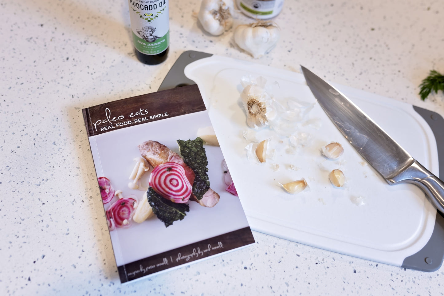 The Paleo Eats Cookbook placed on a cutting board, with bulbs of garlic and a knife, on a plain marble surface.