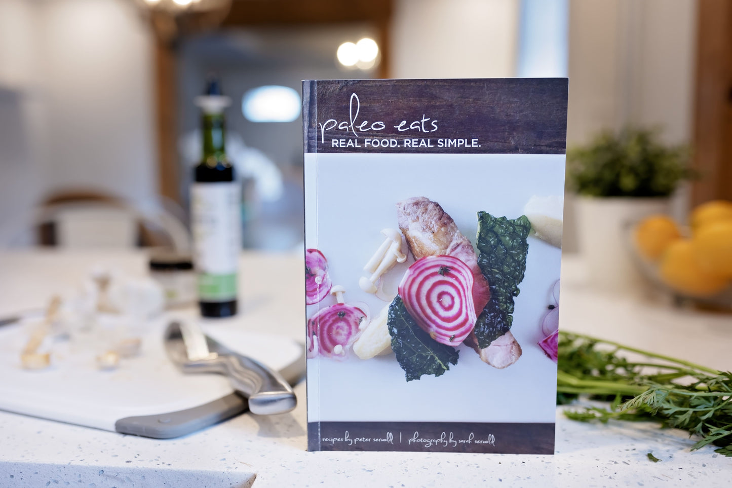 The Paleo Eats Cookbook stands on a kitchen counter, next to a cutting board, a bottle, and some vegetables.