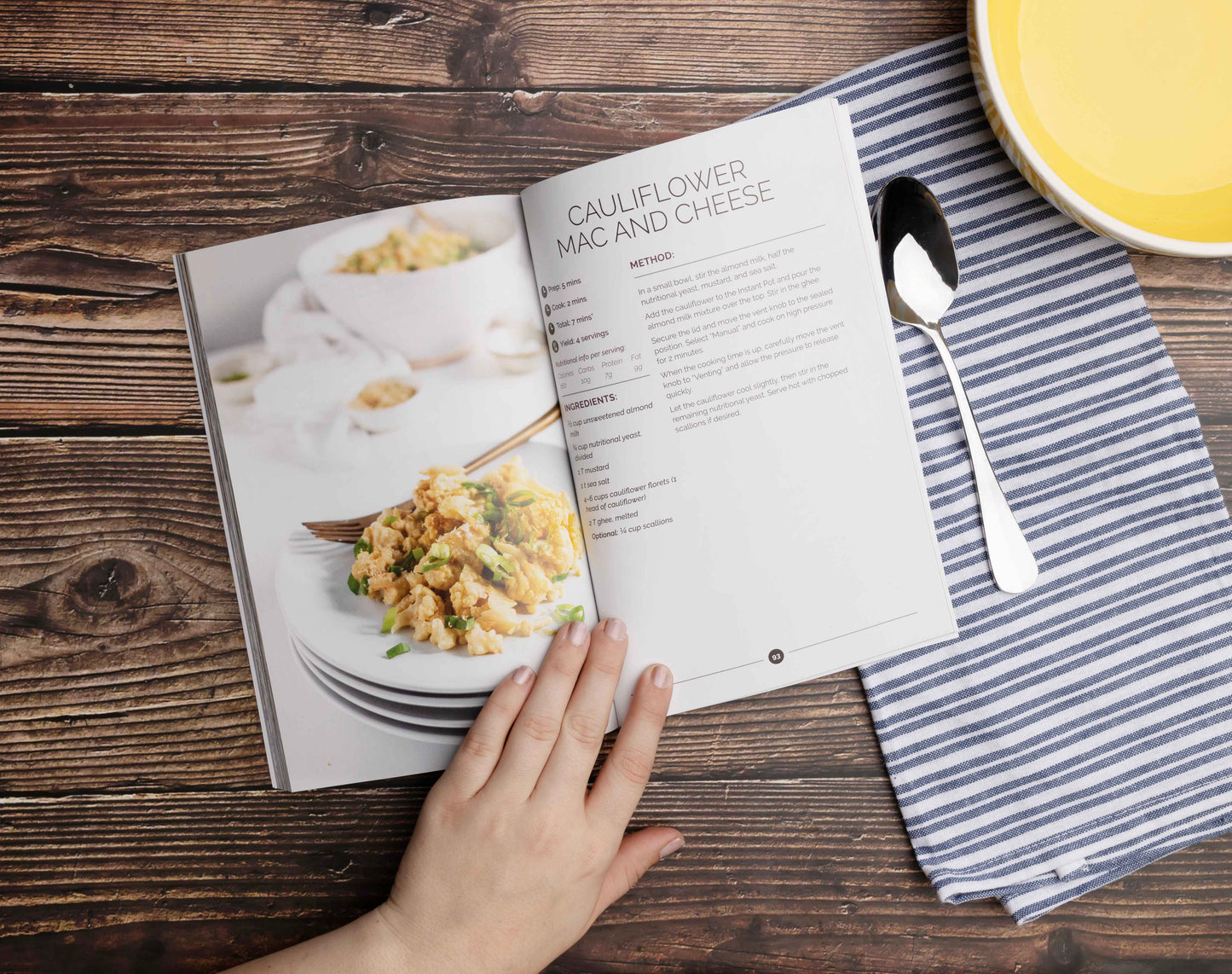 Keto instant pot cookbook opened by a womans's hand on a wooden plain surface. Kitchen cloth and a spoon can be seen.