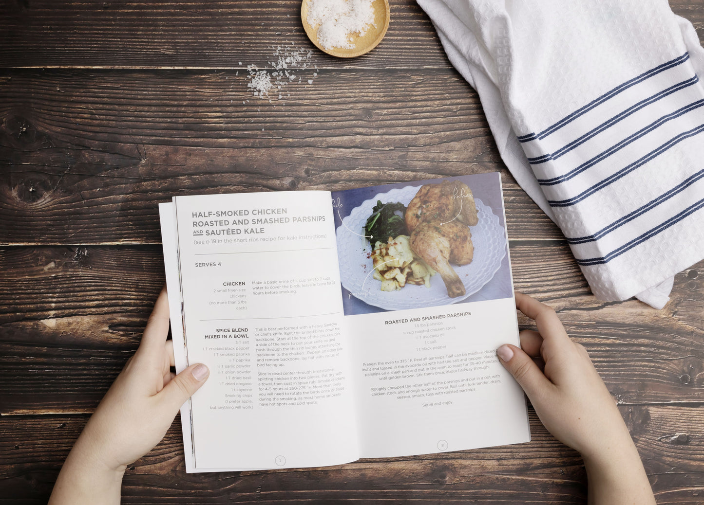 A woman holds an opened Paleo Eats Cookbook on a wooden surface with salt and a kitchen cloth.