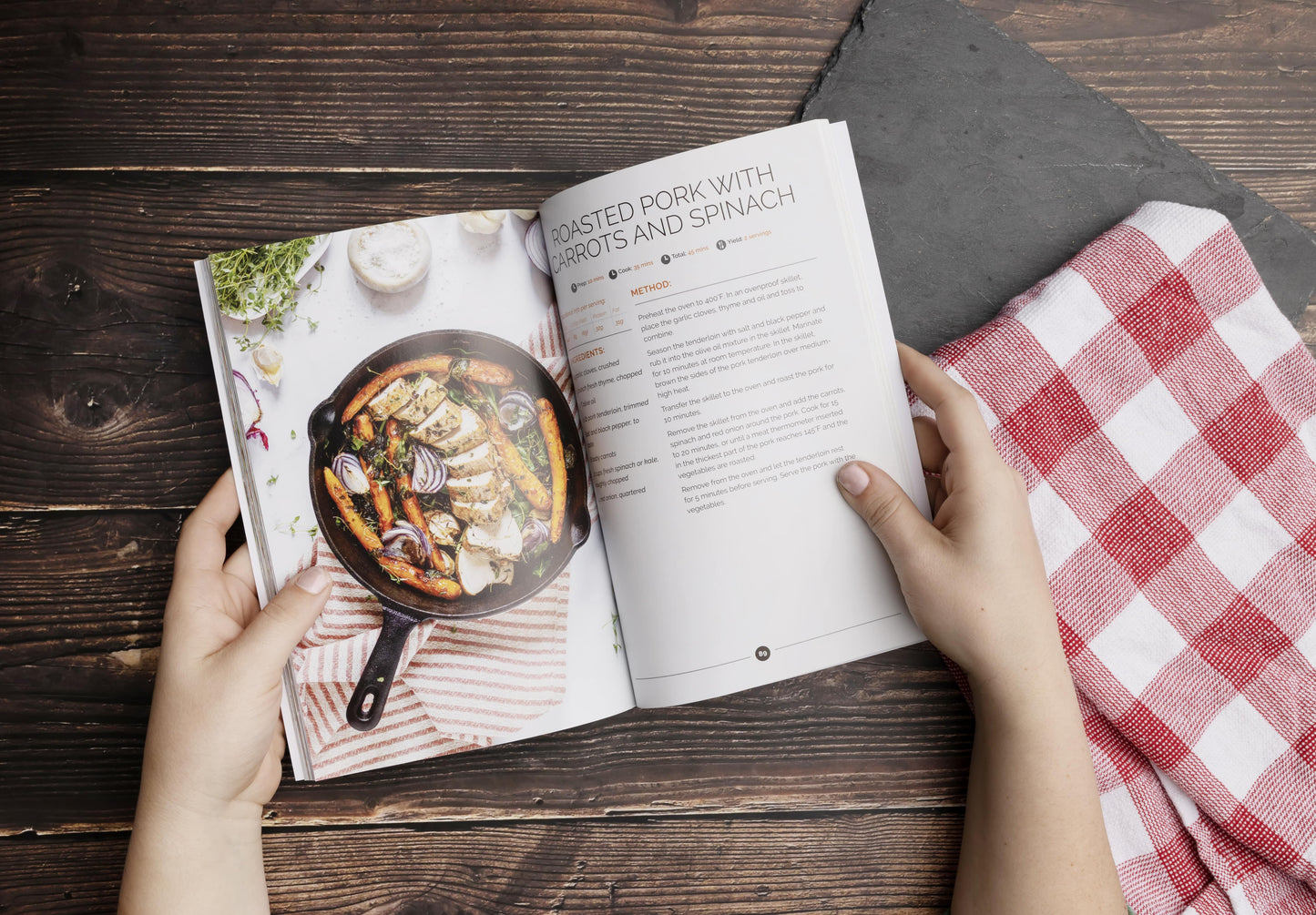 A lady holding an open Keto one-pan cookbook on a wooden surface. A kitchen cloth and cutting board are visible.