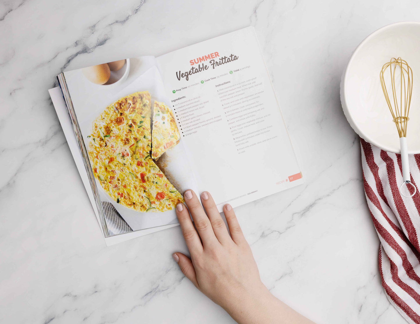A woman's hand opens the Paleo Breakfast Cookbook on a marble surface. A plate with a whisk and a kitchen cloth are placed on the right.