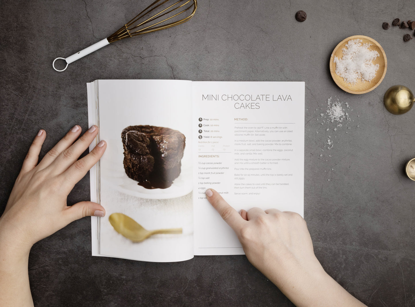 A woman's finger point to a word in an open Keto Sweets Cookbook on a grey surface. A bowl of salt and a whisk can be seen.