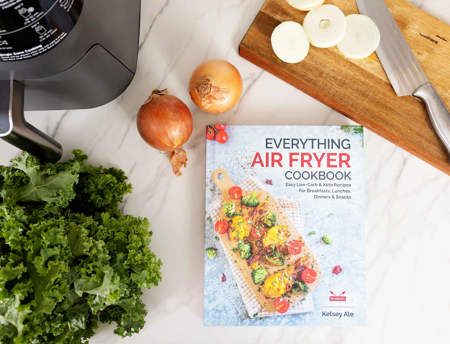 Everything Air Fryer Cookbook laid on a marble surface next to a wooden board containing onions and a knife. Vegetables can be seen on the surface.