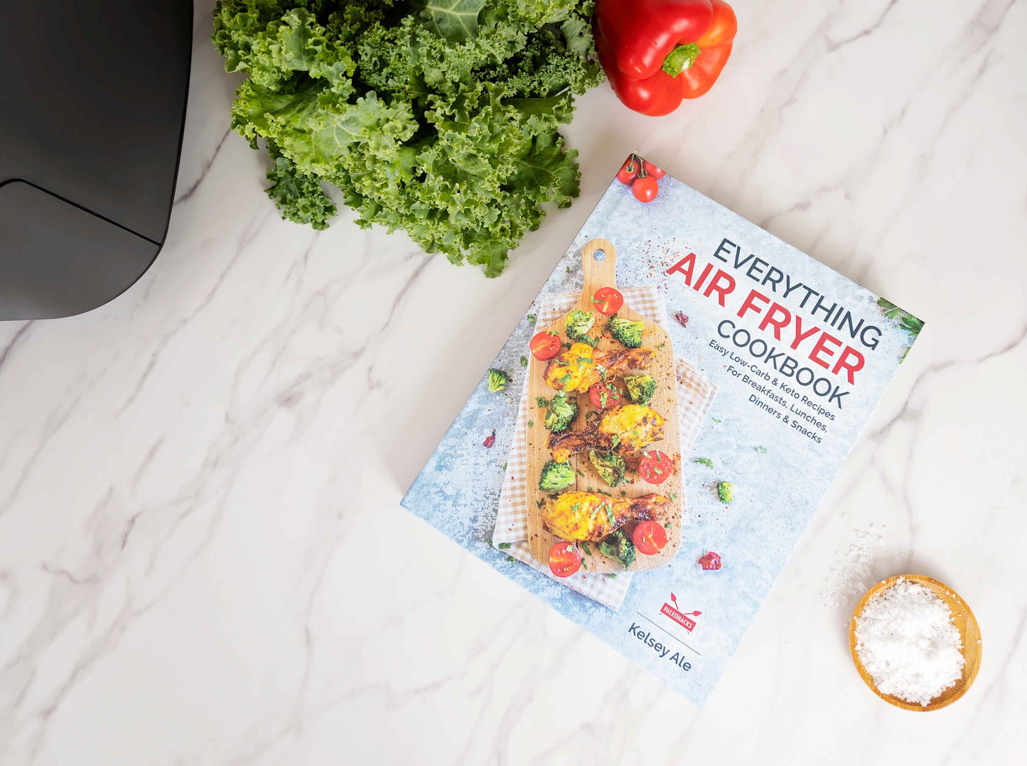 Everything Air Fryer Cookbook laid on a smooth marble surface. Some vegetables can be seen beside it.