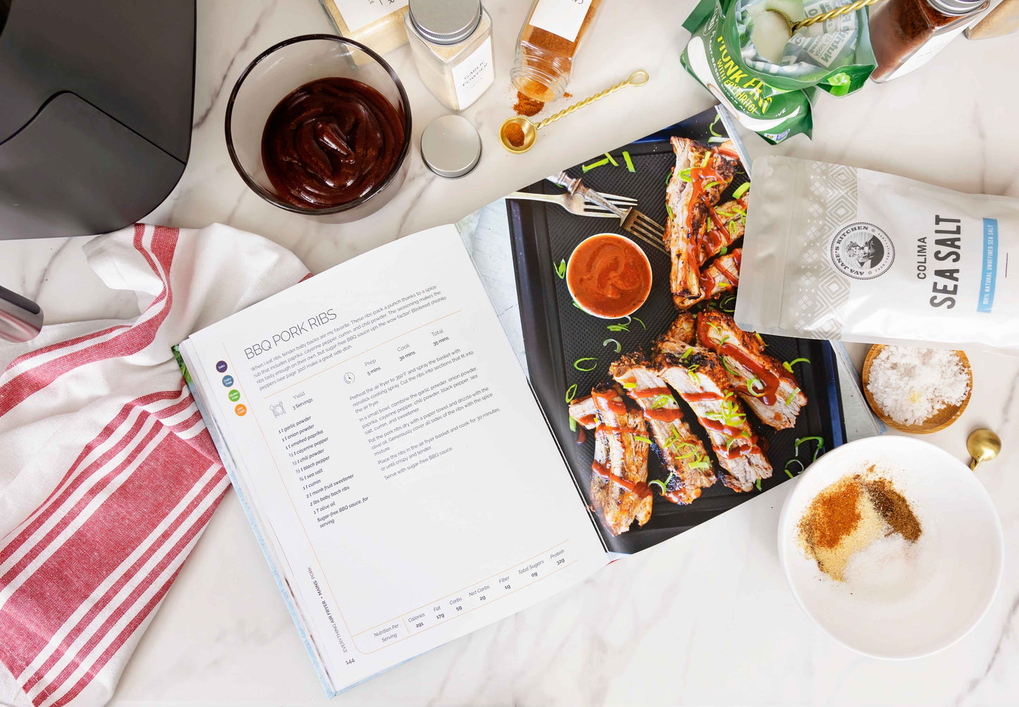 Everything Air Fryer Cookbook opened on a white marble surface, with sea salt, a kitchen cloth, and a couple of other bottled spices.