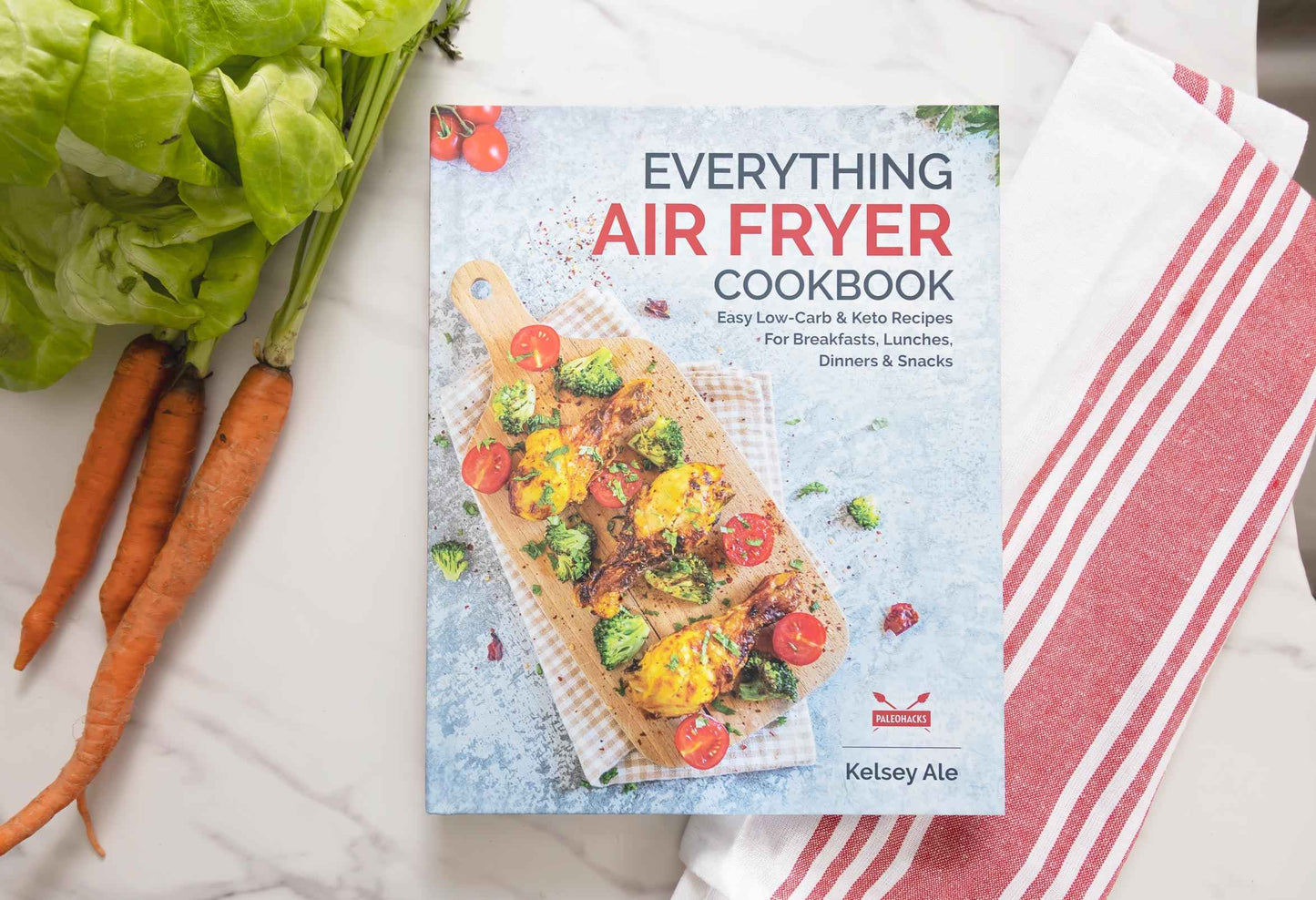 Everything Air Fryer cookbook laid on a marble plain surface . Carrots and kitchen cloth can be seen.