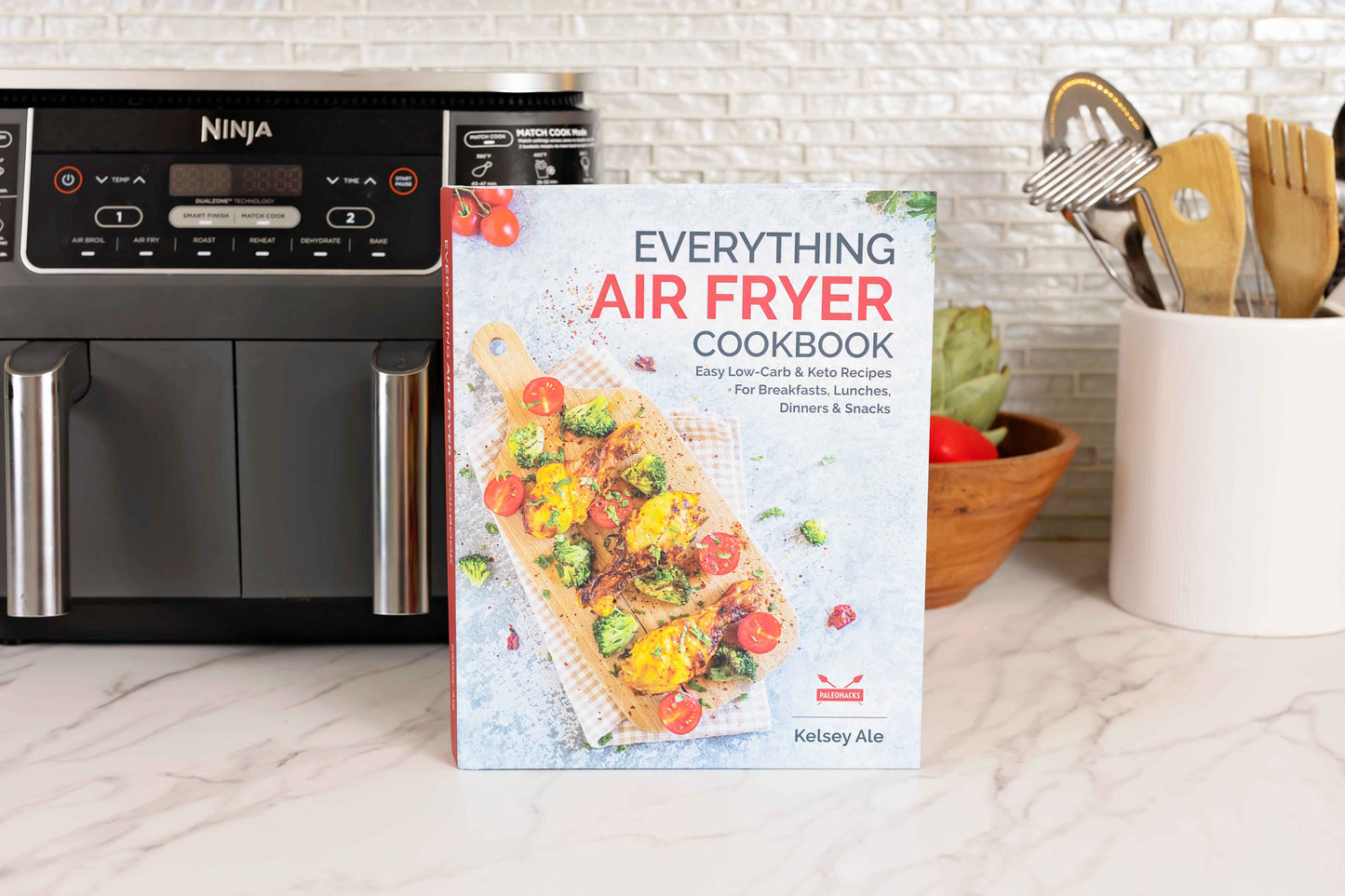 Everything Air Fryer Cookbook leans against an air fryer, accompanied by a bowl of vegetables and a jar of kitchen utensils.