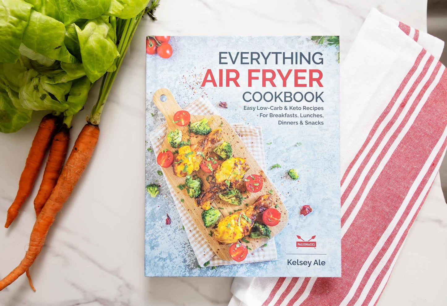 Everything Air Fryer cookbook laid on a marble plain surface . Carrots and kitchen cloth can be seen.