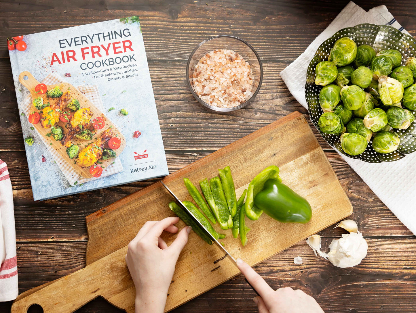 The "Everything Air Fryer" cookbook is placed on a wooden plain surface with a wooden board having sliced vegetables, while a separate bowl of vegetables can be seen.