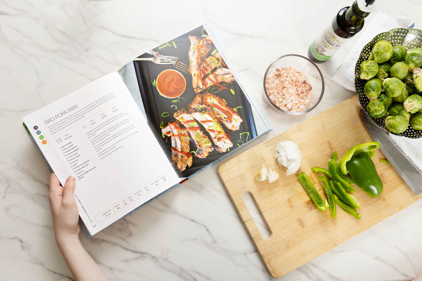 The "Everything Air Fryer" cookbook is open on a marble surface next to a wooden board displaying sliced vegetables. A bowl of vegetables and avocado oil is also visible.