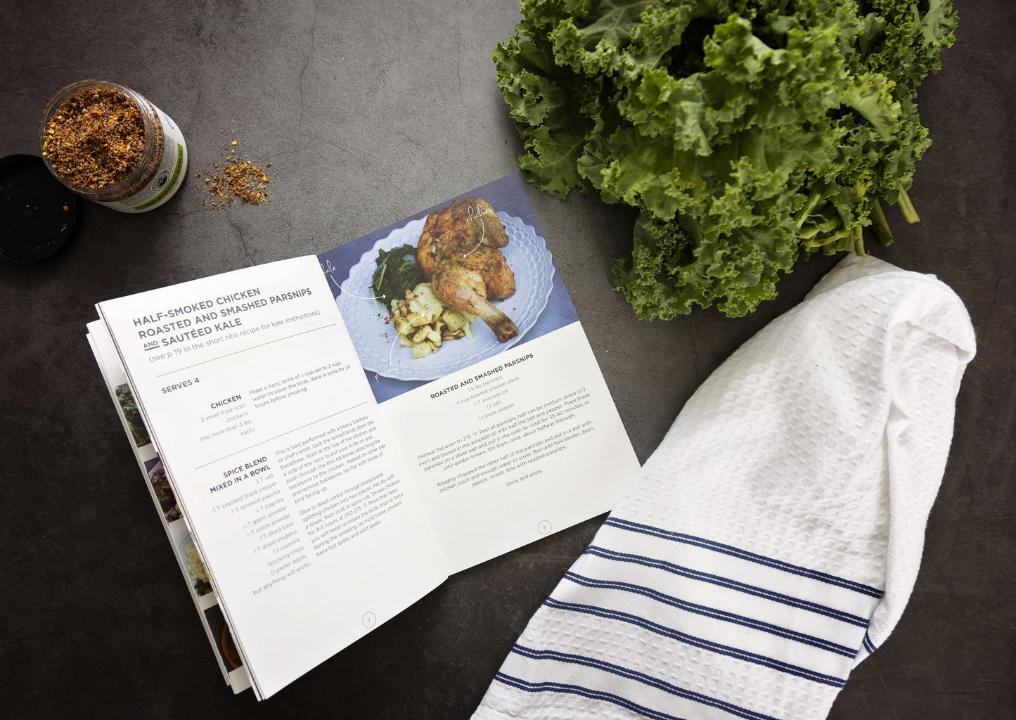 An opened Paleo Eats Cookbook placed on a grey surface with a jar of spices, kale and kitchen cloth.