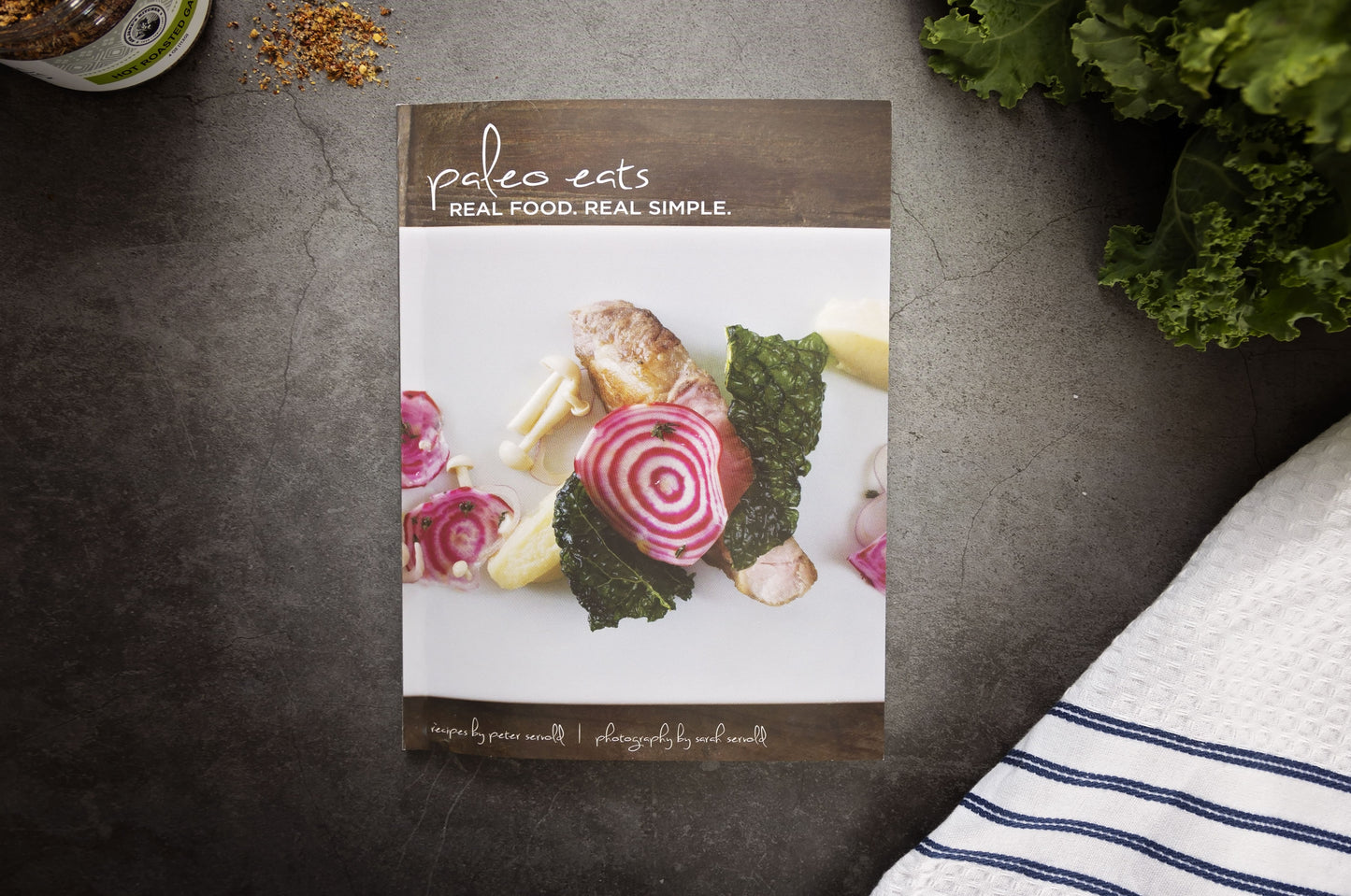 The Paleo Eats Cookbook laid on a grey surface next to a jar of spices. Kale and a kitchen cloth are placed on the right.