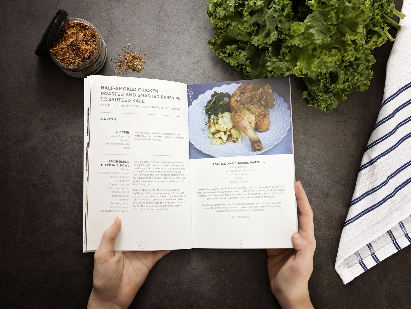 A woman's hand holds an opened Paleo Eats Cookbook over a grey surface. A jar of spices, kale, and a kitchen cloth are visible around.