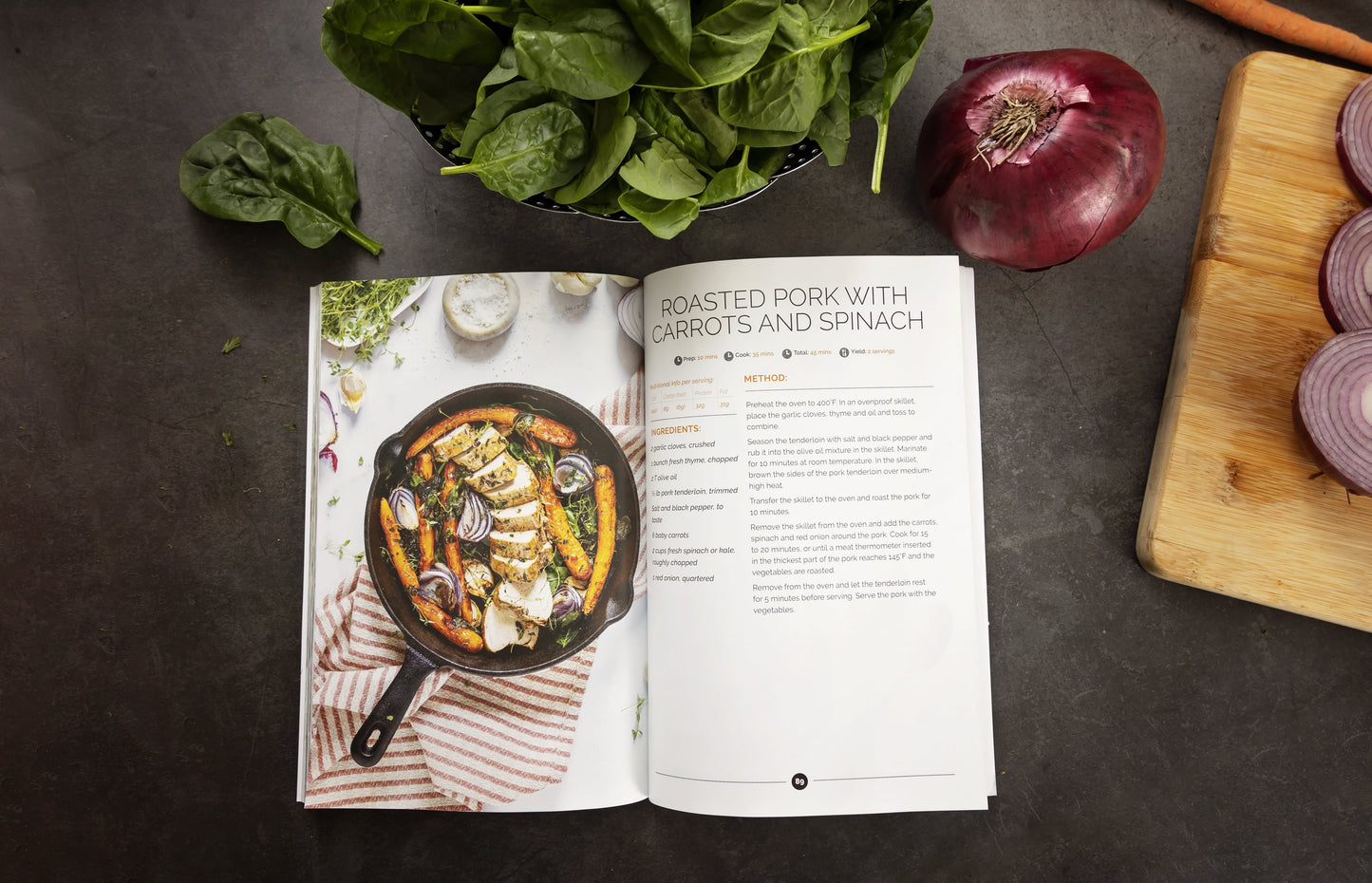 An open Keto one-pan cookbook laid on a grey surface, accompanied by a bowl of vegetables, an onion, and a few sliced onions on a cutting board.