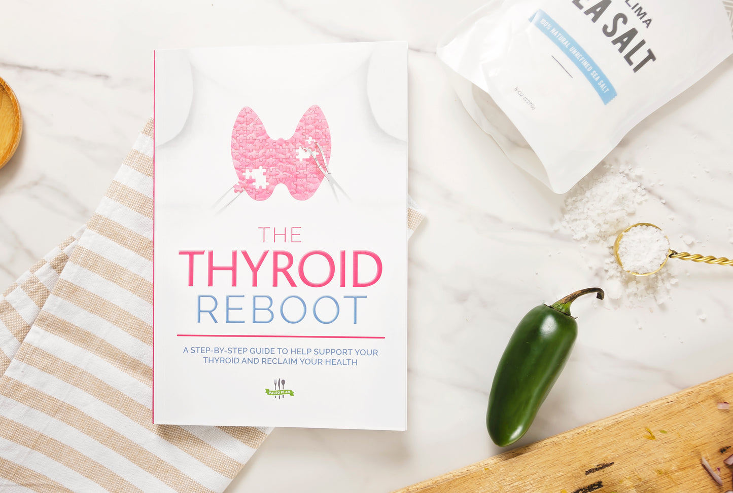 The Thyroid Reboot Book laid on a white plain surface, next to a kitchen cloth, a pack of salt, and a jalapeno pepper.
