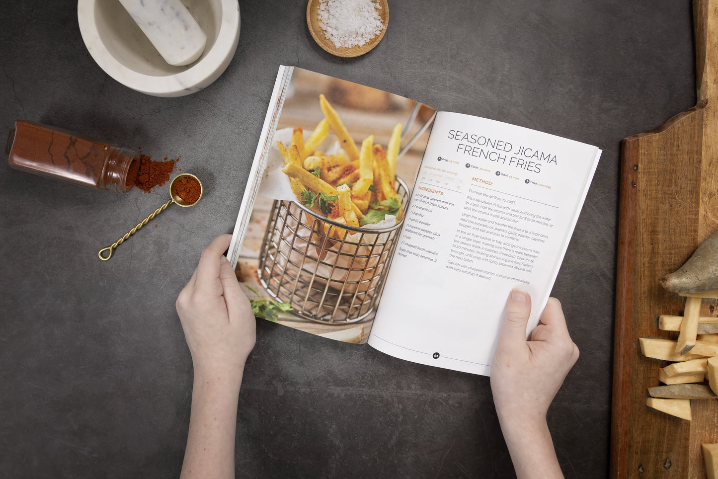 A woman holds an open Keto Air Fryer cookbook. Spice bottles, salt, a wooden board, and other items are visible.