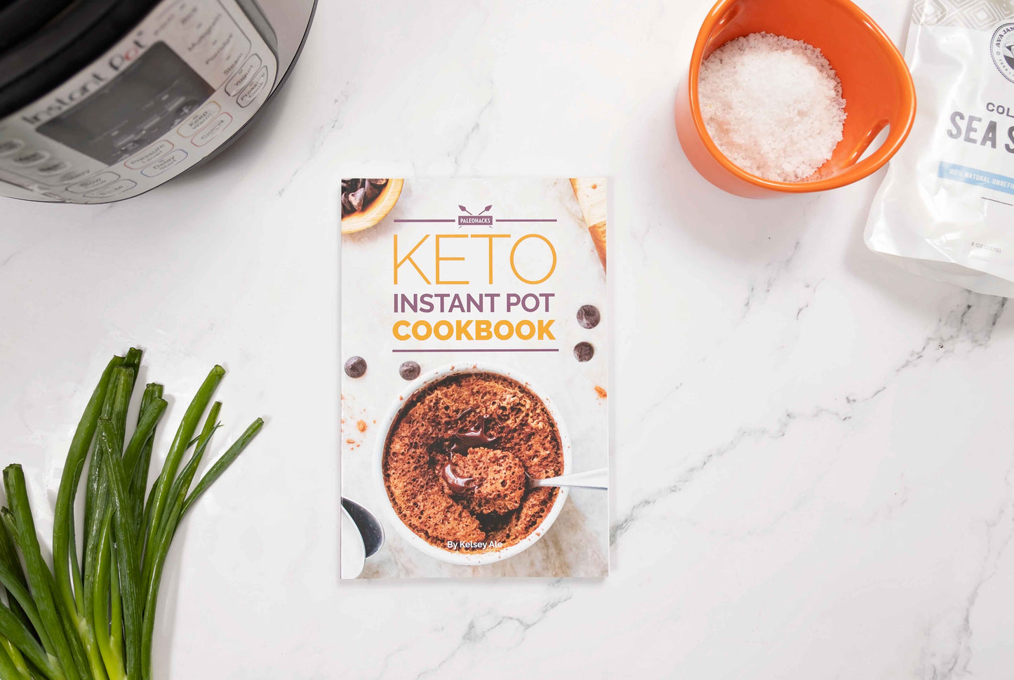 Keto instant pot cookbook laid on a marble surface. An instant pot, a bowl of sea salt and spring onions can be seen.