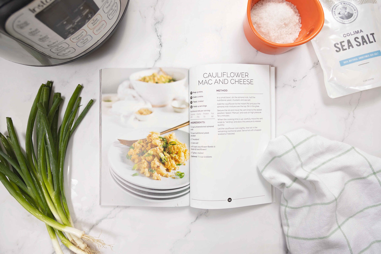 An opened Keto Instant Pot Cookbook laid on a marble surface. Spring onions, sea salt and a kitchen cloth can be seen.