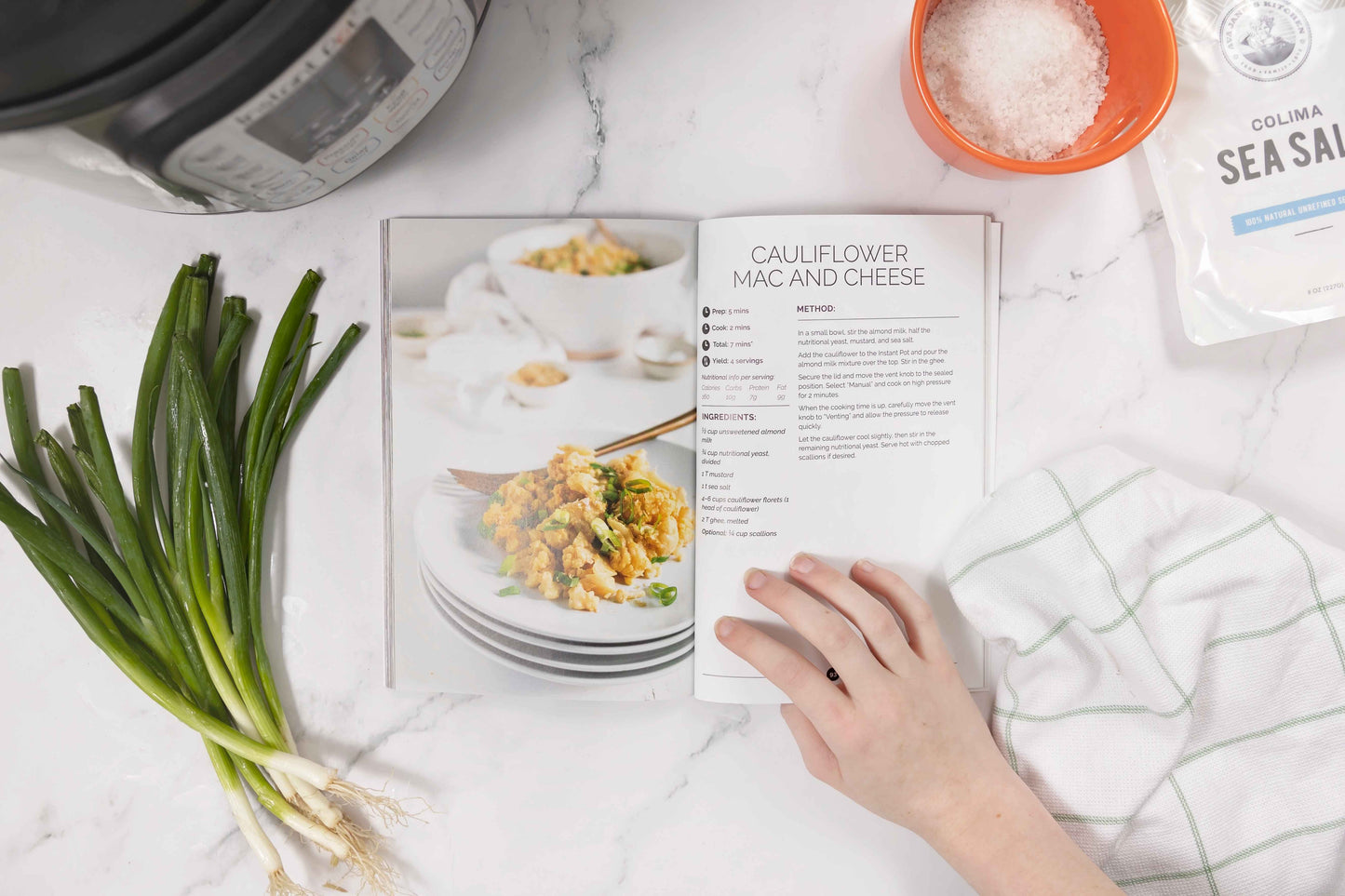 A woman's hand opens a page of the Keto Instant Pot Cookbook on a marble surface. Sprigs of fresh spring onions, a bowl of salt, and kitchen cloth are all visible.