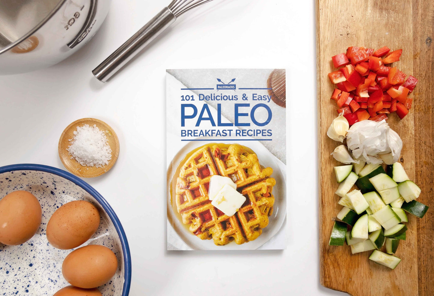 The Paleo Breakfast Recipes Cookbook placed on a plain white surface, accompanied by a bowl of eggs, sea salt, and a cutting board with diced vegetables.