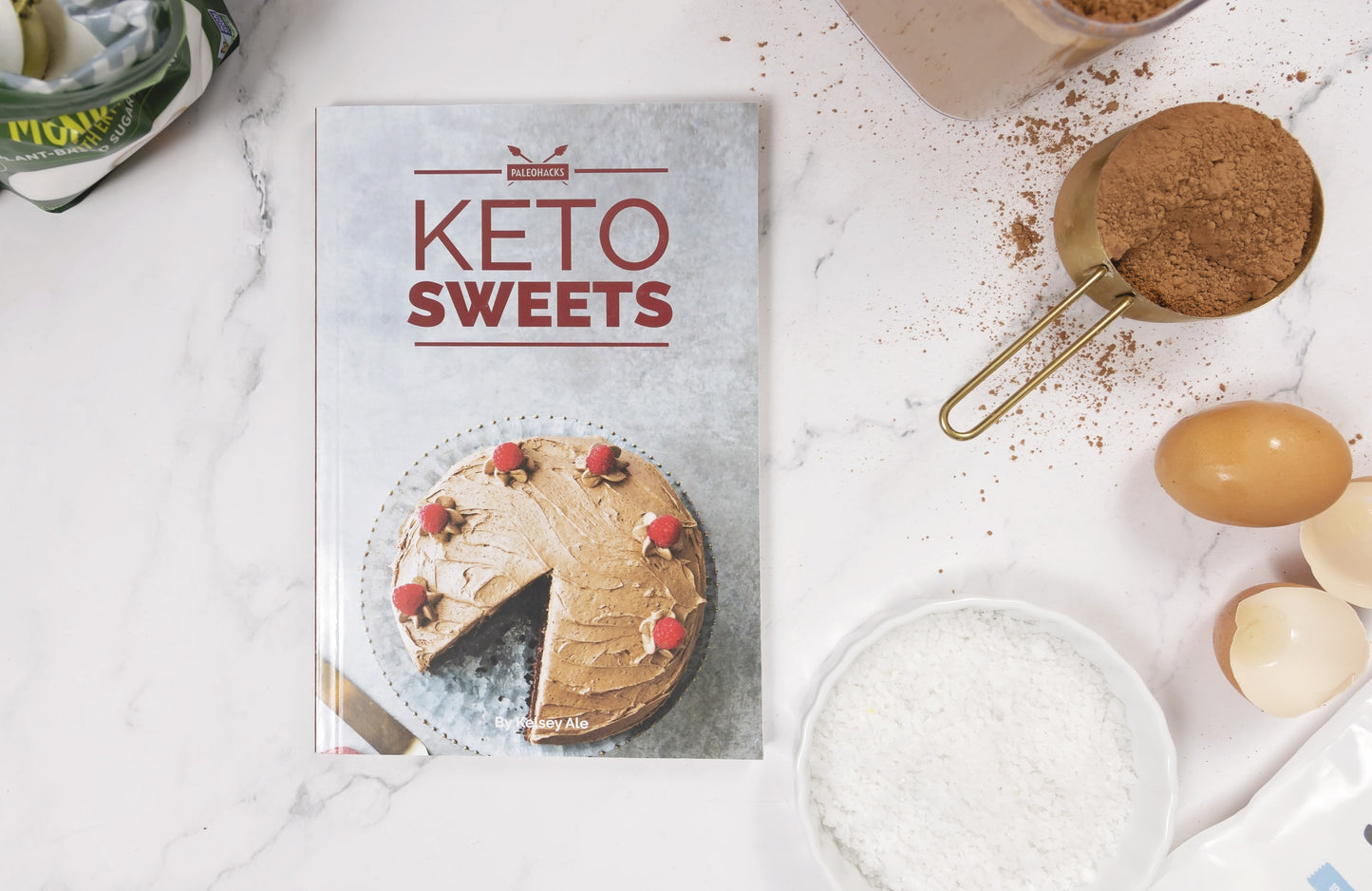 The Keto Sweets Cookbook placed on a white marble surface. A bowl of flour, cacao powder, and eggs can be seen on the right.