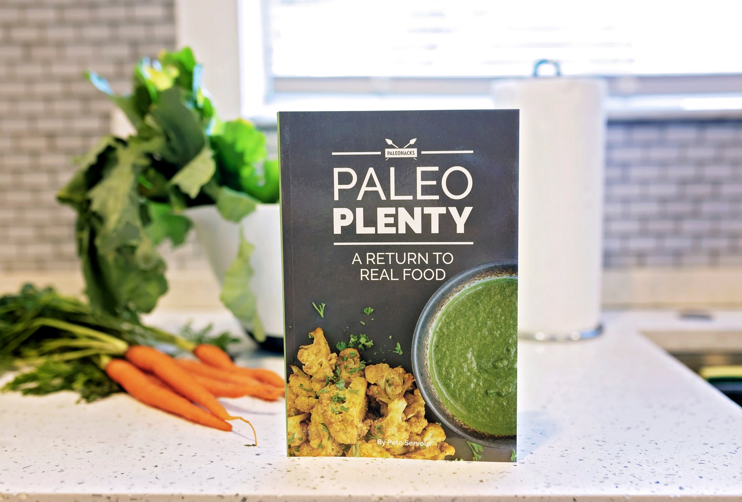 The Paleo Plenty Cookbook stands on the marble kitchen counter. Carrots and a bowl of vegetables can be seen in the background.