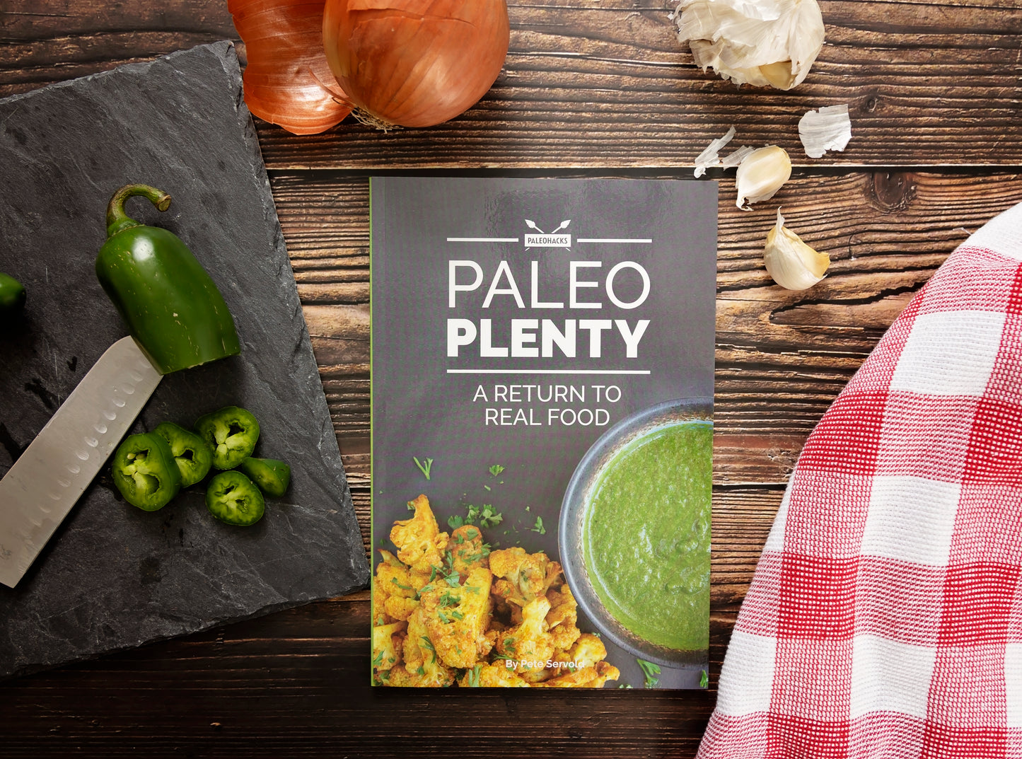 The Paleo Plenty Cookbook placed on a plain wooden surface, surrounded by chopped peppers on a board, onions, garlic, and a kitchen cloth.