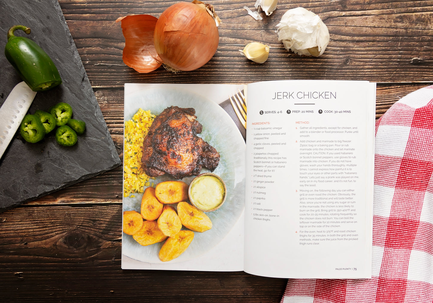 An opened Paleo Plenty Cookbook laid on a wooden surface next to a chopping board with chopped peppers. Onions, garlic, and a kitchen cloth can also be seen.