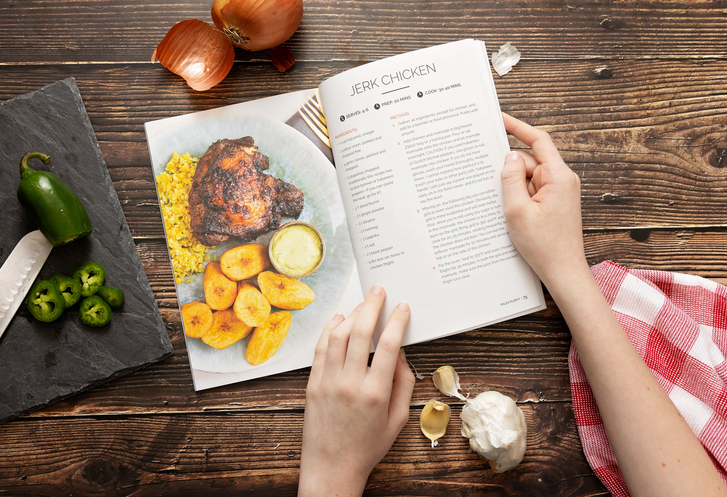 A woman's hands holds an opened Paleo Plenty Cookbook on a plain wooden surface, beside a cutting board with chopped peppers. Onions, garlic, and a kitchen cloth.
