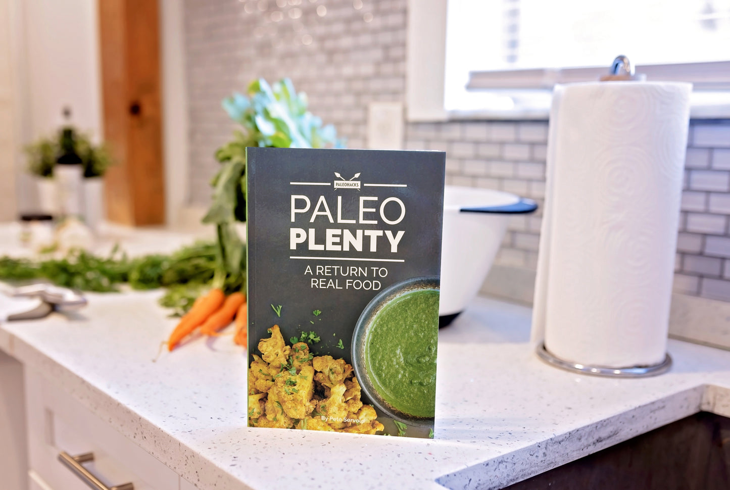 The Paleo Plenty Cookbook stands on the marble kitchen counter. Carrots, a bowl of vegetables and a roll of disposable paper can be seen in the background.