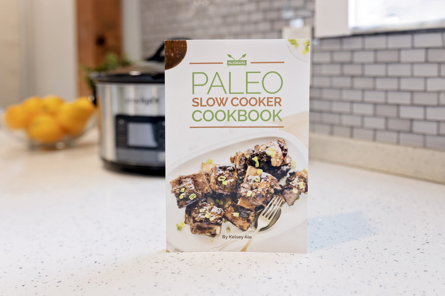The Paleo Slow Cooker Cookbook stands on a marble kitchen counter. A pot and a bowl of fruit are seen in the background.