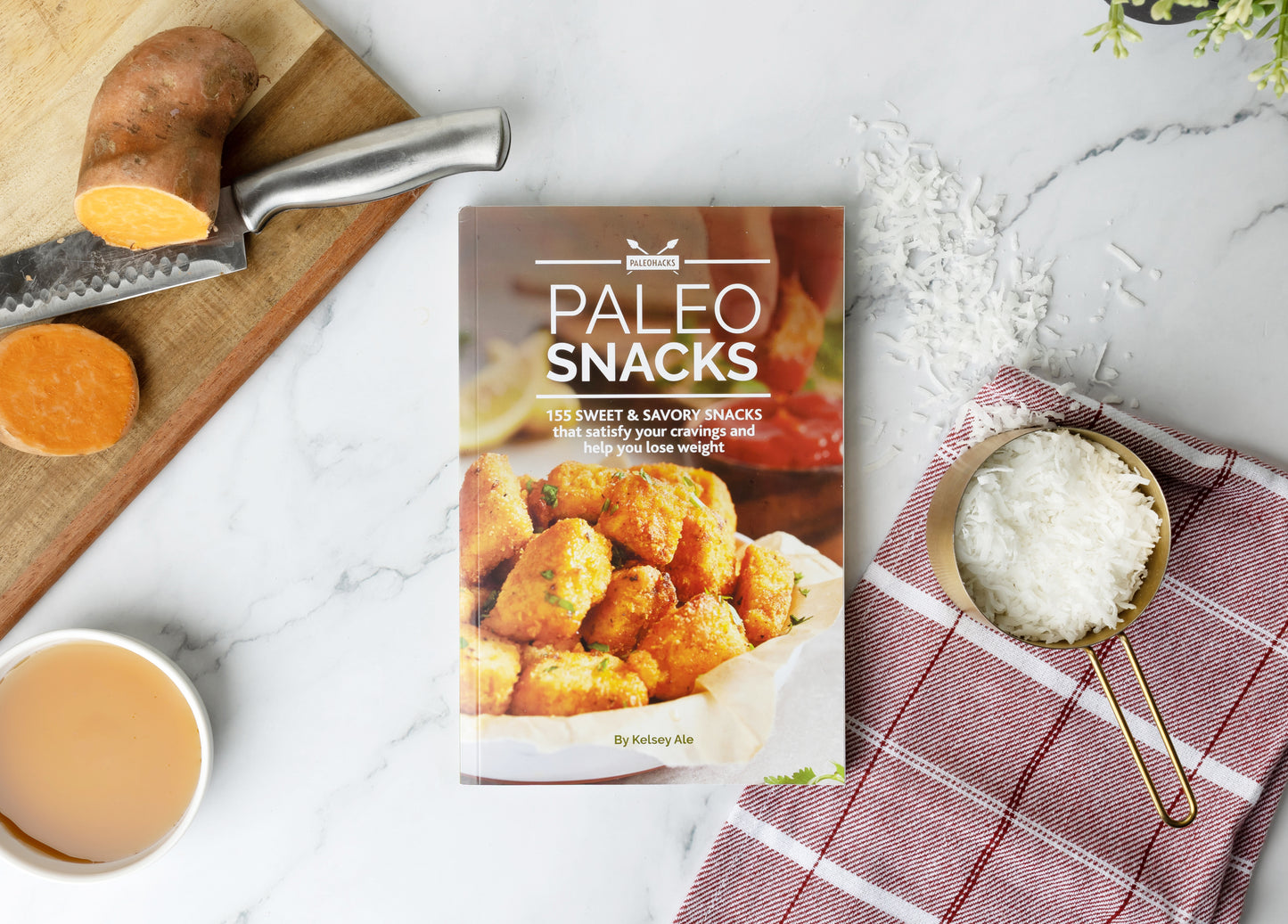 Paleo Snacks Cookbook laid on a marble surface next to a cutting board with a knife and sweet potatoes. Kitchen cloth and shredded coconut can be seen on the right.