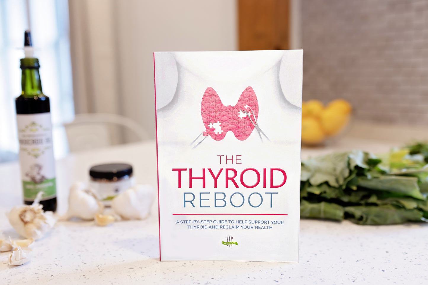 The Thyroid Reboot book stands on a plain marble surface. Bottles of avocado oil, bulbs of garlic, and vegetables can be seen in the background.
