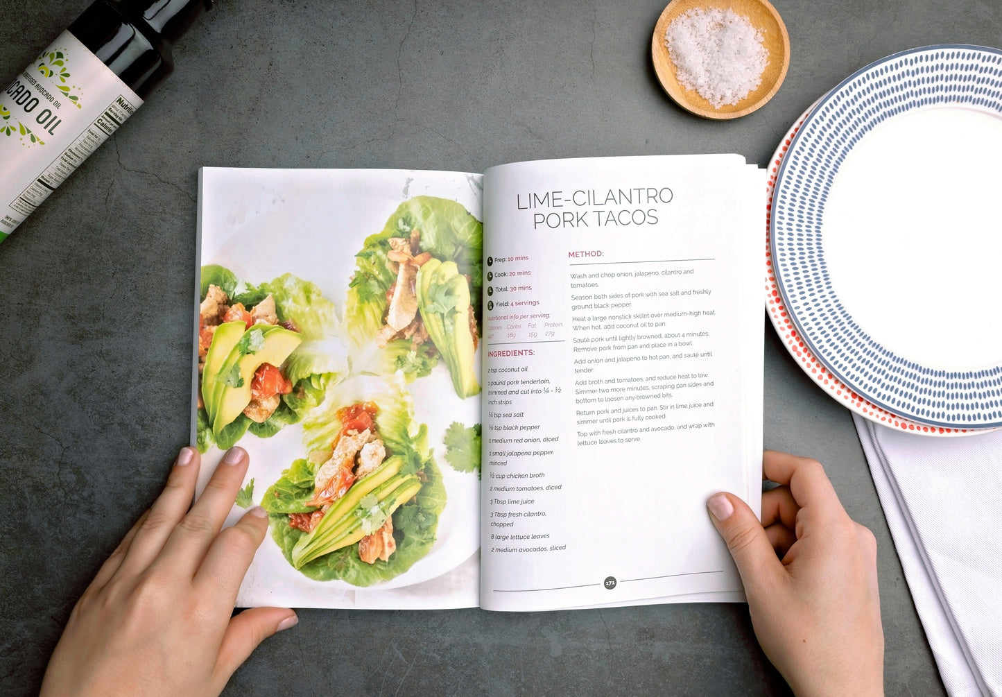 A woman holds an opened Thyroid Reboot book on a grey plain surface. A bottle, sea salt and plates can also be seen.