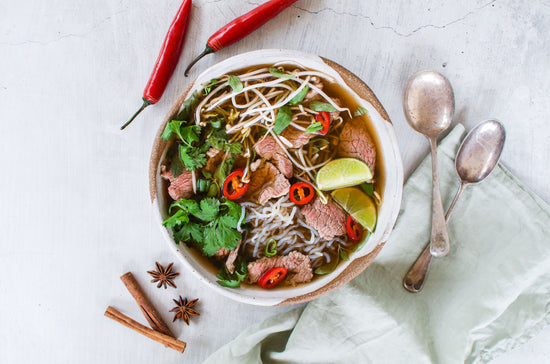 A bowl of Beef Brisket Pho placed on a marble plain surface. Chili pepper and spoons are visible in the scene.