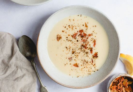 A bowl of Creamy Cauliflower Soup with Bacon and a spoon placed on a white plain surface.