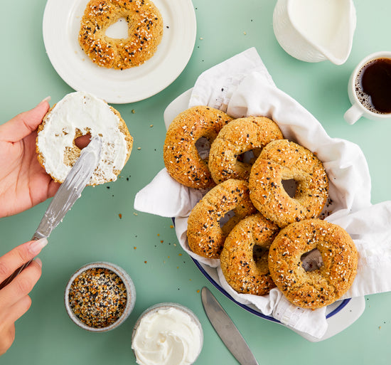  A bowl of bagels with everything seasoning. A jar of cream and seasoning, a knife and tea cups can be seen.