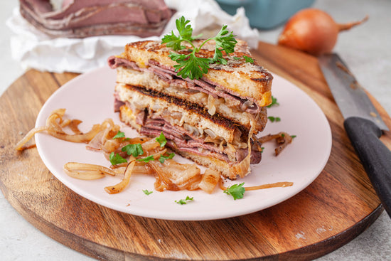 A white plate of grilled pastrami with cheese on a wooden board.