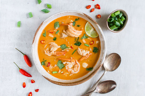 A bowl of Keto Tom Yum placed on a white marble surface with pepper, vegetables and spoons.