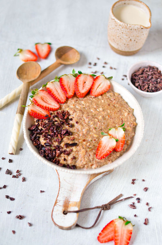 A bowl of Paleo porridge on a wooden board placed on a white surface with strawberries, chocolates and a jar of cream.
