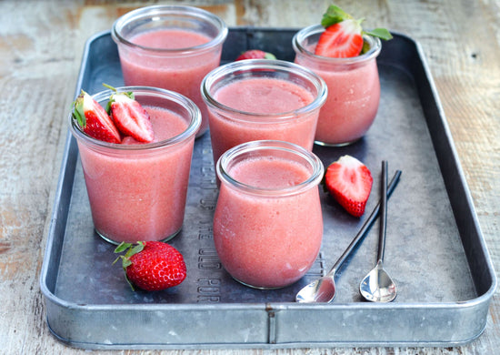 Five glassess of strawberry jello with spoons, placed in a grey tray on a wooden surface.