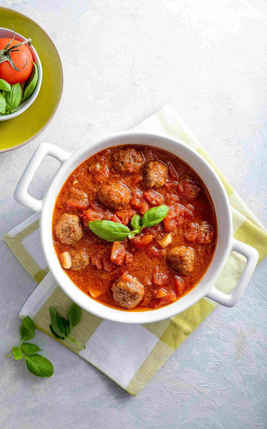 A white bowl of meatballs in a tomato sauce on a white surface with a kitchen cloth.