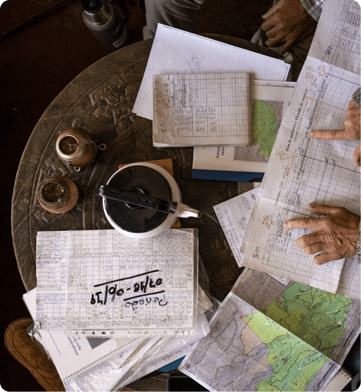 Wrinkled hands sifted through a collection of maps and spreadsheets, on an aged table with two metallic pots and a bowl, steeped in antiquity.