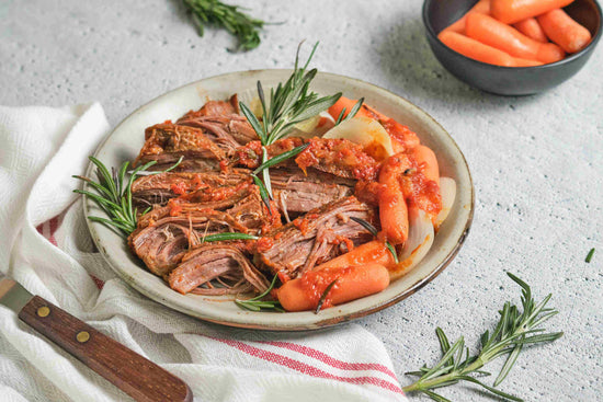 A plate of Pot Roast with Baby Carrots on a marble plain surface. A bowl of carrots and some herbs can be seen.