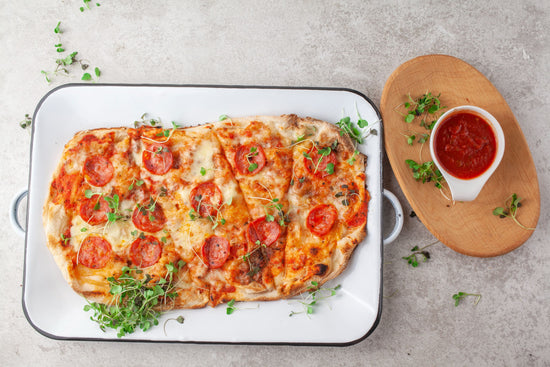 A white tray of flat bread pizza with a sauce bowl on a wooden board.