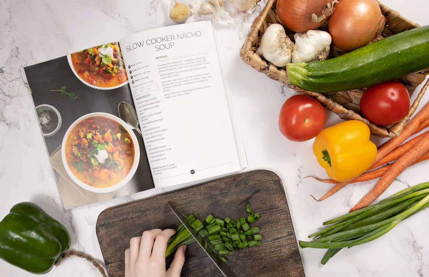 An open cookbook, some vegetables sliced with a knife on a wooden board and a basket of vegetables on the right all laid on a grey marble plain surface.