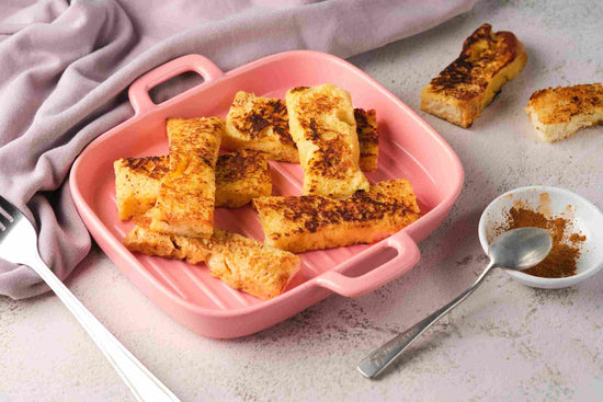 A pink plate of French toast on a plain surface. A bowl of spice with a spoon can be seen.